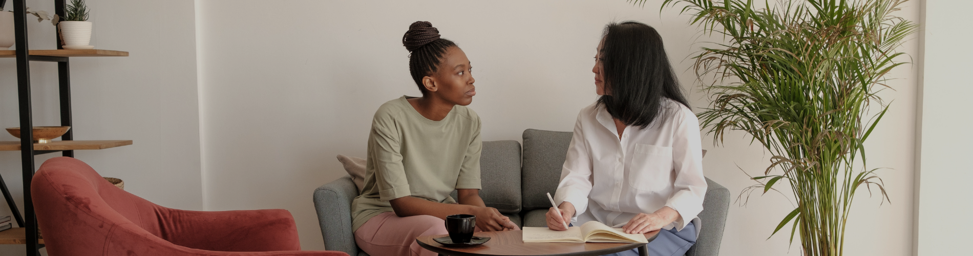 Two women sitting on a couch. One is writing in a notebook on the table in front of them.